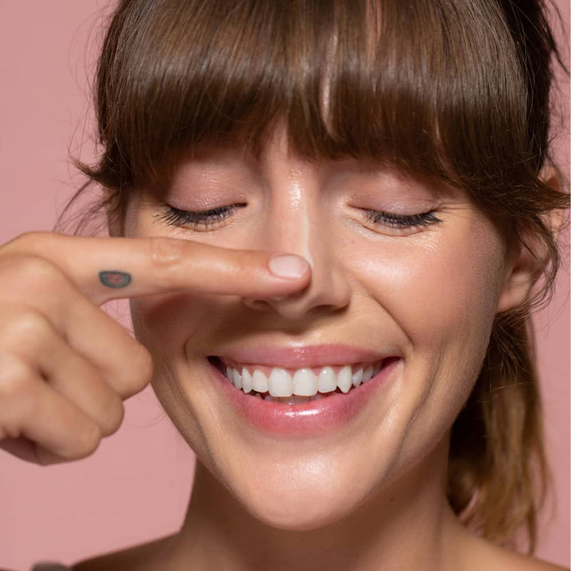 Close up of a six gldn model looking down and smiling and applying the Essential Moisturizer down her nose and on one finger she has a cute tattoo all against a pink background