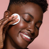 Close up face photo of a Six Gldn model with one hand to her face using the Botanical Toner with a cotton pad on her cheek. She is looking down and to the side as she smiles with a light pink background